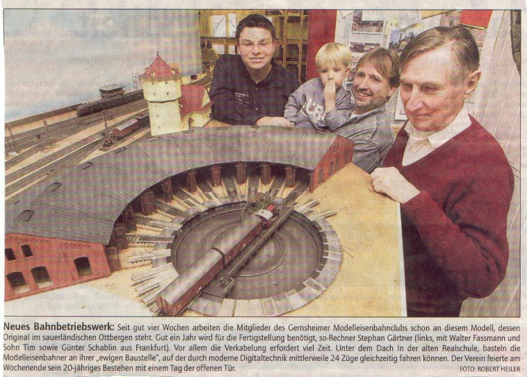 Historischer Pressefotoausschnitt: Vier Personen, darunter ein Kind, stehen hinter einem im Bau befindlichen Modellbahnbetriebswerk mit großer Lokomotivdrehscheibe und Ringlokschuppen.