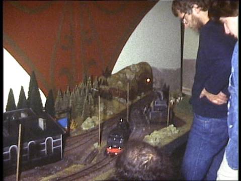 Ein Modellzug fährt durch die fertig aufgebaute Modellbahnlandschaft mit Bergen, Tunneln und Bäumen. Ein Vereinsmitglied beobachtet den Fahrbetrieb.