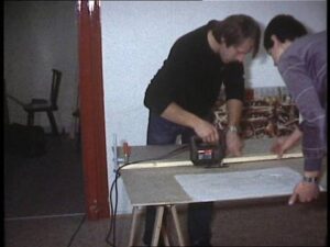 Zwei Vereinsmitglieder bearbeiten eine Holzplatte mit einer elektrischen Stichsäge, um Bauteile für die Modellbahnanlage zuzuschneiden.