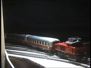 Eine rote Diesellokomotive zieht einen rot-beigen Personenwagen über die Gleise der Modellbahnanlage bei der ersten Probefahrt am 22. November 1986.