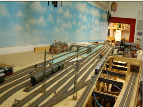 Blick über den linken Teil der MEC-Modellbahnanlage mit zahlreichen parallel verlaufenden Gleisen, Oberleitungsmasten, Zügen und Bahnhofsgebäuden. Im Hintergrund ist eine blau bemalte Himmelskulisse zu sehen.