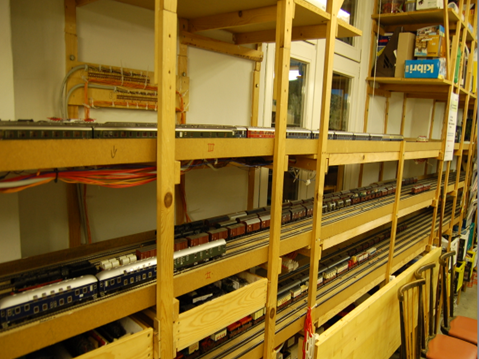 Mehrstöckige Holzkonstruktion der Modellbahnanlage im zweiten Clubraum. Auf mehreren Ebenen verlaufen Gleise mit abgestellten Personen- und Güterzügen; darunter sind Kabel und Leitungen sichtbar.