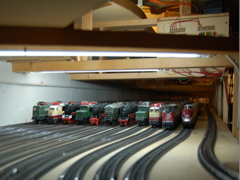 Blick in den Schattenbahnhof des MEC Gernsheim. Auf zahlreichen parallelen Gleisen stehen verschiedene Modelllokomotiven, darunter Dampflokomotiven, Diesellokomotiven und elektrische Lokomotiven. Über den Gleisen sind Beleuchtung und Verkabelung zu sehen.