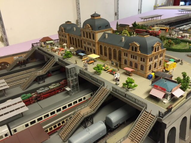 Ausgestaltetes Empfangsgebäude des Schöfferstadt Hauptbahnhofs mit belebtem Vorplatz, Treppen und Aufzug zu den tiefer liegenden Bahnsteigen.