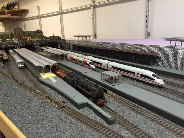 Blick auf die Gleise und Bahnsteige des Schöfferstadt Hauptbahnhofs mit Dampflokomotive, ICE-Zügen und mehreren Bahnsteigdächern.