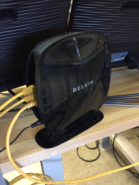 Schwarzer Belkin-WLAN-Router mit angeschlossenen Netzwerkkabeln auf einem Holzregal vor zwei Monitoren.