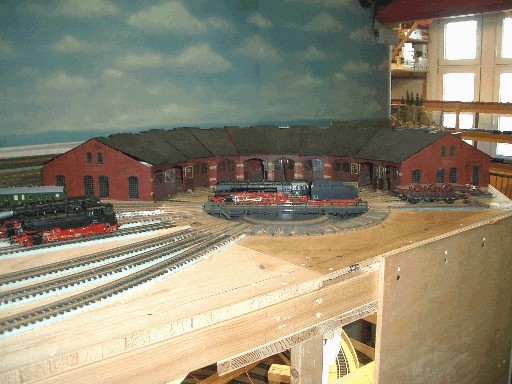 Historische Aufnahme des noch im Bau befindlichen Bahnbetriebswerks mit einer Drehscheibe, einer Dampflokomotive und einem großen roten Ringlokschuppen. Die Gleise liegen teilweise noch auf einer ungestalteten Holzplatte.