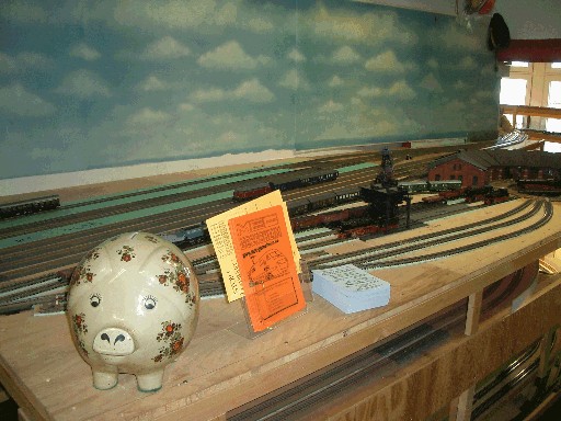 Weißes Sparschwein mit Blumenmuster steht neben einem orangefarbenen Informationsschild am Rand der Modellbahnanlage. Dahinter sind Gleise und Lokomotiven des Bahnbetriebswerks zu sehen.