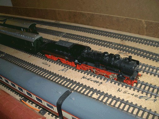 Schwarze Dampflokomotive der Baureihe P 8 mit rotem Fahrwerk und angehängtem Wagen auf einem Abstellgleis
