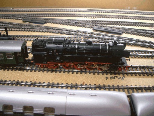 Schwarze Tenderdampflokomotive der Baureihe 65 mit rotem Fahrwerk auf einem Gleis neben abgestellten Reisezugwagen