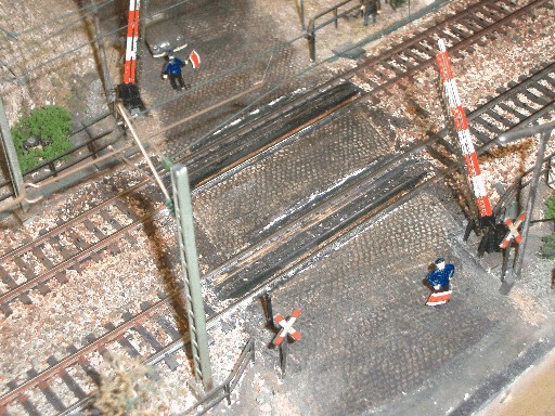 Modellbahn-Bahnübergang aus der Vogelperspektive mit zwei geöffneten rot-weißen Schranken und kleinen Figuren