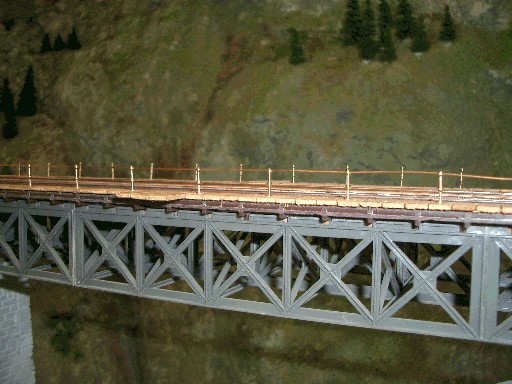 Seitliche Ansicht einer grauen Modellbahn-Fachwerkbrücke mit goldfarbenen Geländerpfosten und dünnen, durchgezogenen Drähten