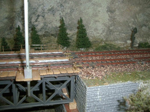 Rechtes Ende der Modellbahn-Fachwerkbrücke mit gespannten Geländerdrähten, Oberleitungsmast und gemauertem Widerlager