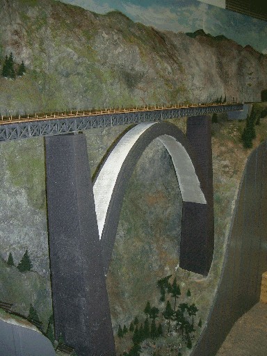Lange graue Modellbahnbrücke mit Fachwerkträger und hohem gemauertem Bogen über einer felsigen Schlucht