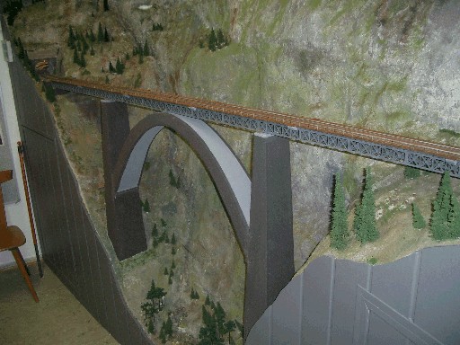 Lange Modellbahnbrücke mit grauem Bogen und Fachwerkträgern vor einer felsigen Gebirgslandschaft