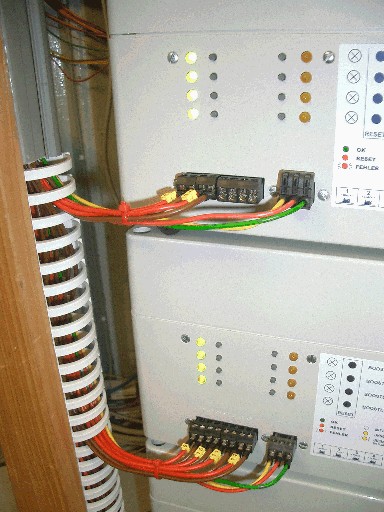 Detailaufnahme der Anschlüsse zweier Booster mit roten, grünen und gelben Kabeln, Steckverbindungen und leuchtenden Kontrollanzeigen.