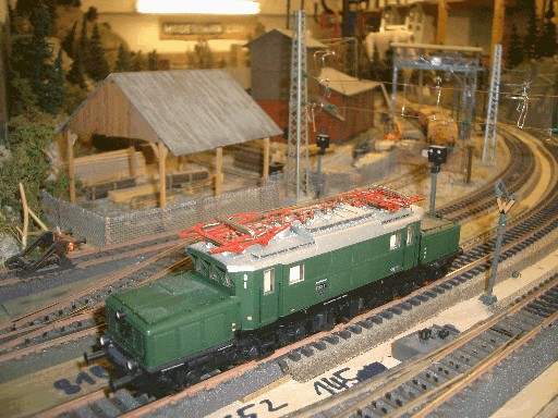 Grüne Elektrolokomotive der Baureihe E93 auf der Modellbahnanlage vor einem Sägewerk mit Holzlager, Oberleitungsmasten und mehreren Gleisen.