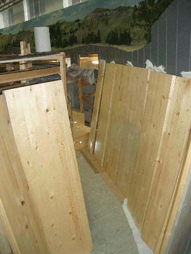 Mehrere Holzbretter und größere Holzplatten stehen zum Streichen aufrecht im Clubraum. Im Hintergrund ist die Gebirgslandschaft der Modellbahnanlage zu sehen.
