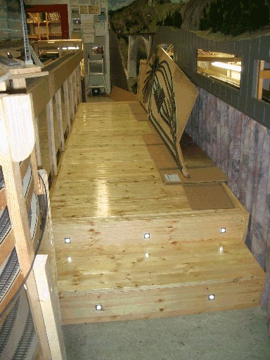 Fertig lackiertes Besucherpodest aus hellen Holzbrettern neben der Modellbahnanlage. Zwei breite Stufen mit eingelassenen Leuchten führen auf die erhöhte Plattform.