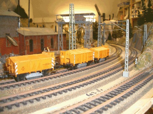 Mehrere gelbe Reinigungswagen stehen auf einem gebogenen Gleis der Modellbahnanlage. Im Hintergrund sind ein rotes Gebäude und Oberleitungsmasten zu sehen. Die Lokomotive ist nicht im Bild.