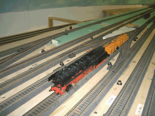 Schwarze Dampflokomotive mit rotem Fahrwerk zieht mehrere gelbe Reinigungswagen durch den noch im Bau befindlichen Hauptbahnhof. Zahlreiche parallel verlaufende Gleise sind auf der ungestalteten Grundplatte verlegt.