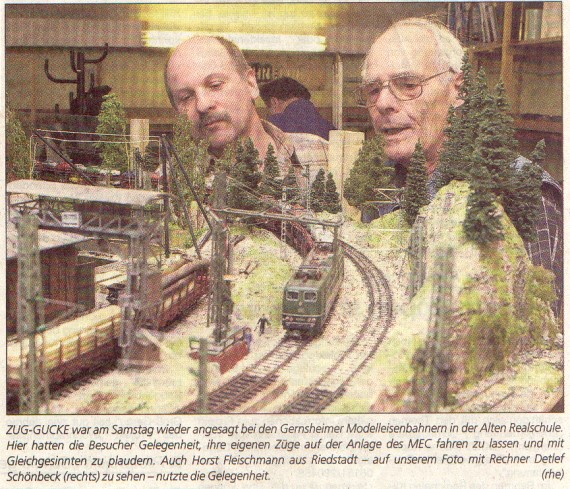 Historischer Pressefotoausschnitt: Zwei Männer betrachten eine Modellbahnlandschaft mit elektrischen Lokomotiven, Gleisen, Oberleitungen, Brücken und bewaldeten Hügeln.