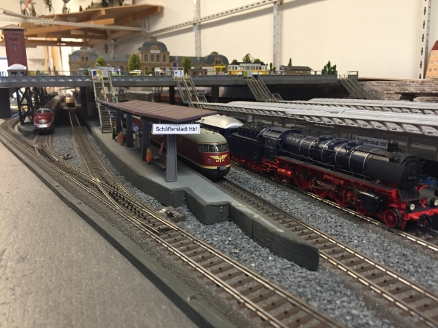 Modellbahnhof Schöfferstadt Hbf mit mehreren Bahnsteigen, einer schwarzen Dampflokomotive und zwei roten Triebwagen. Fußgängerbrücken verbinden die Bahnsteige. Sichtbare Eisenbahnsignale fehlen.