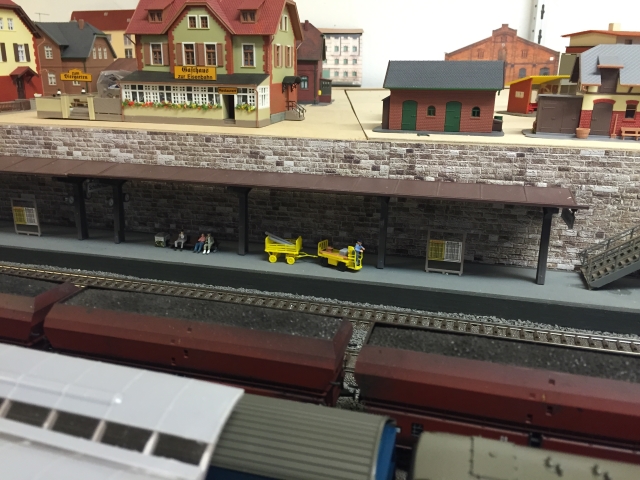 Bahnsteig mit Ankunft-/Abfahrtstafel, Gepäckwagen sowie Reisenden auf einer Bank