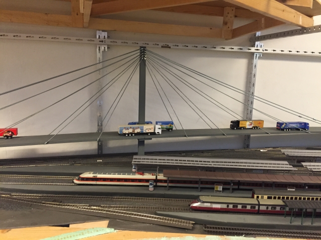 Modell einer Schrägseilbrücke mit Lastwagen über den Gleisen des Hauptbahnhofs Schöfferstadt. Darunter stehen mehrere Züge an den Bahnsteigen.