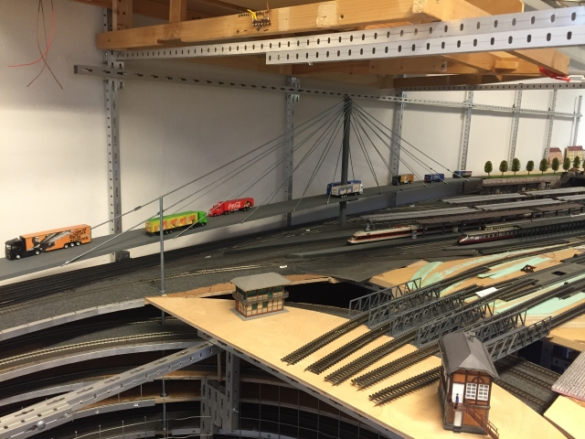 Seitliche Ansicht der Schrägseilbrücke mit mehreren Lastwagen. Die Straße steigt über eine Rampe an und überquert die Gleise der Modellbahnanlage.
