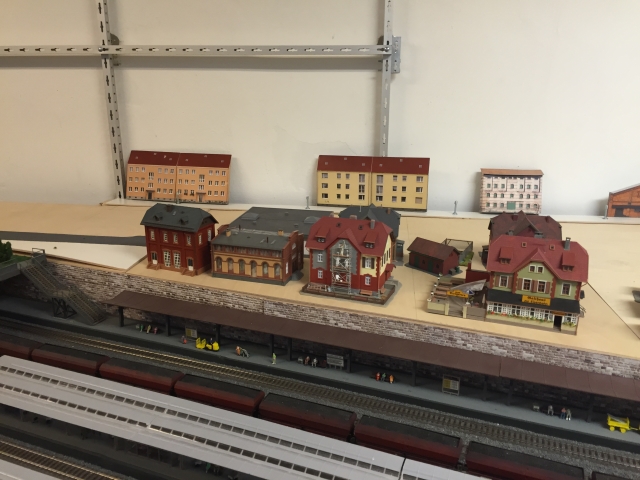 Weitere Ansicht der Stellprobe mit mehreren Modellhäusern auf der noch kahlen Stadtfläche. Darunter sind die Bahnsteige und abgestellte Züge zu sehen.