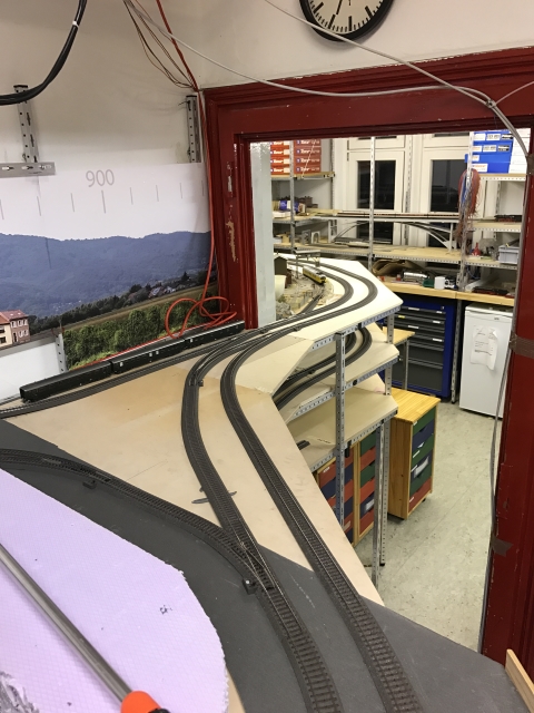 Mehrere gebogene Modellbahngleise führen auf einer erhöhten Trasse durch eine Türöffnung in den hinteren Teil des Clubraums.