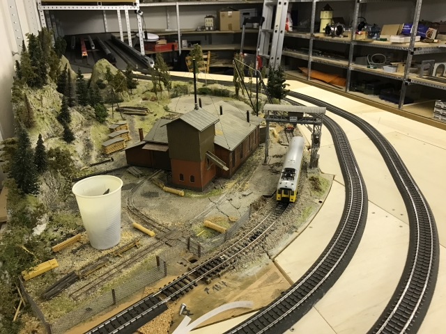 Modell eines Sägewerks mit mehreren Gebäuden, Holzlager und Gleisanschluss. Eine gebogene Bahnstrecke führt um das Sägewerksgelände herum.