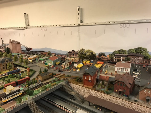 Mittlerer Bereich der Modellstadt Schöfferstadt mit Wohn- und Gewerbegebäuden, Straßen, einer Straßenbahn und einer Brücke über den Bahngleisen. Im Hintergrund sind Industrieanlagen und eine Landschaftskulisse zu erkennen.