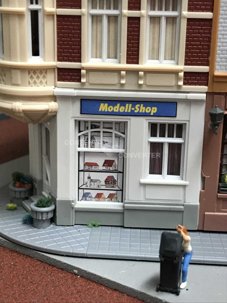 Ein Frau schaut in eine offene Mülltonne hinein. Dahinter eine Laden-/Häuserzeile mit dem Modell-Shop.