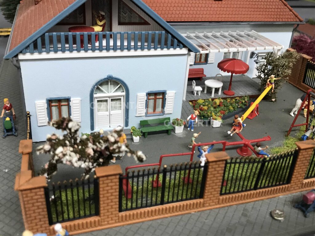 Kindergartengebäude sowie spielende Kinder auf der umzäunten Freifläche beim Wippen und Schaukeln