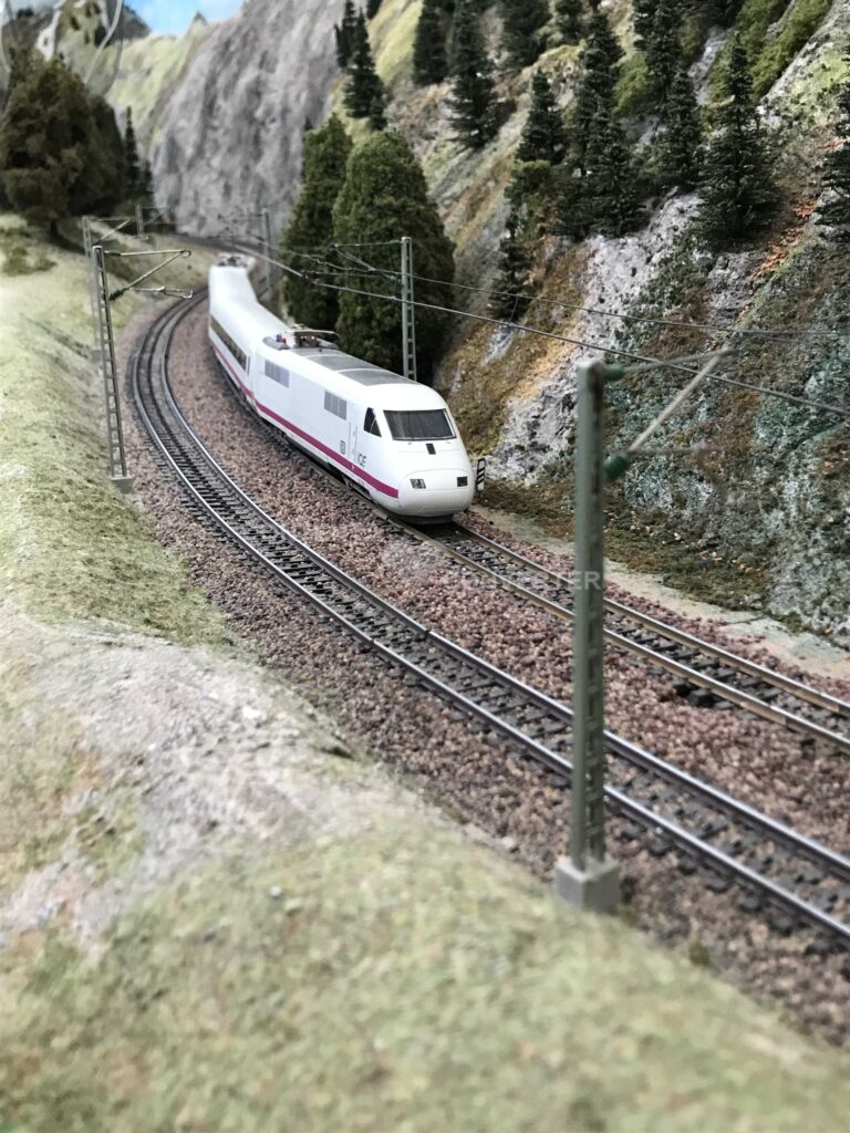 Weißer ICExperimental mit rotem Zierstreifen auf einer zweigleisigen, elektrifizierten Modellbahnstrecke, die in einer Kurve durch eine felsige, bewaldete Gebirgslandschaft führt.