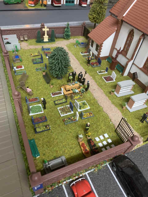 Friedhof neben der Kirche in Schöfferstadt mit kleinen Gräbern, Urnengräbern und einem Weg. Mehrere Modellfiguren haben sich zu einer Beerdigung am offenen Grab versammelt.