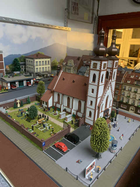 Kirche mit zwei Zwiebeltürmen und angrenzendem Friedhof auf der Modellbahnanlage des MEC Gernsheim. Zwischen den Gräbern verlaufen kleine Wege, vor der Kirche liegt ein Platz mit Figuren und parkenden Autos.