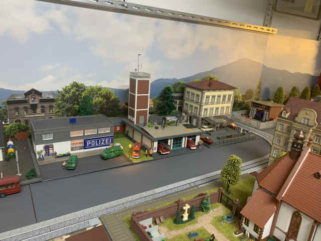 Modellbahnszene in Schöfferstadt mit Polizeistation, Feuerwehrgerätehaus und mehreren Einsatzfahrzeugen. Im Hintergrund stehen weitere Gebäude, im Vordergrund ist ein Teil des Friedhofs zu sehen.