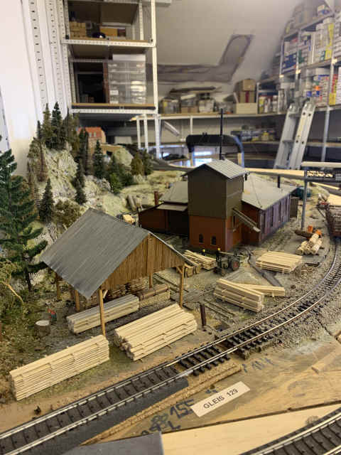 Vorderansicht eines Sägewerks auf der Modellbahnanlage des MEC Gernsheim mit Holzgebäuden, gestapelten Brettern und einem überdachten Holzlager. Im Vordergrund verläuft ein Bahngleis, dahinter erhebt sich ein bewaldeter Felshang.