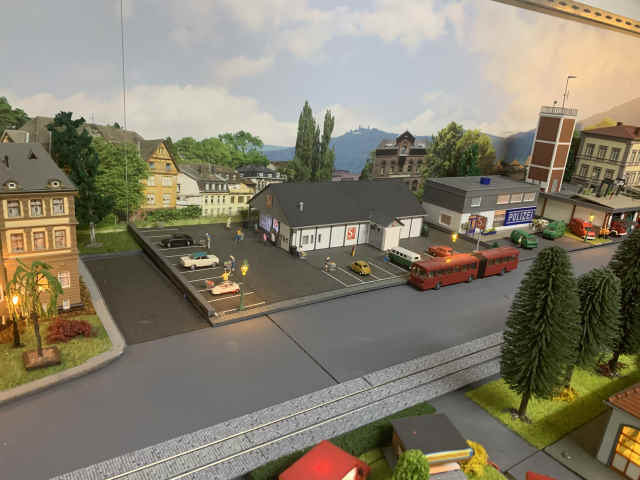 Supermarkt mit Parkplatz und mehreren Autos in der Modellstadt Schöfferstadt. Vor dem Gebäude fährt eine rote Straßenbahn, rechts sind die Polizeistation und die Feuerwehr zu sehen.