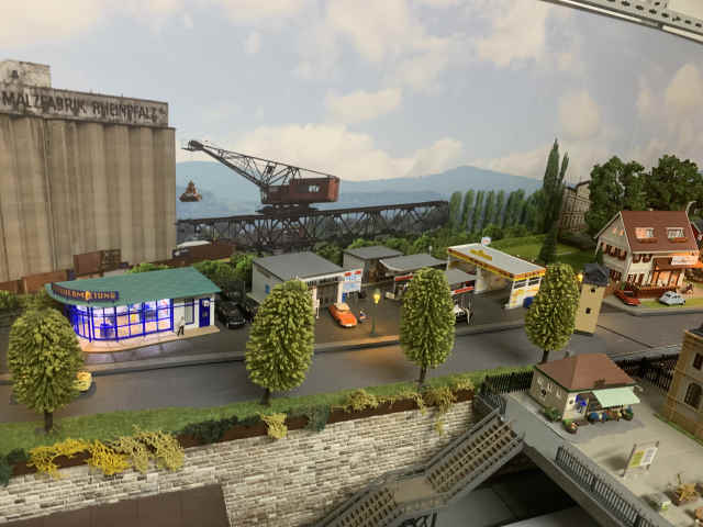 Modellbahnszene mit Tankstelle, Autowaschanlage und beleuchtetem Gebäude einer Autovermietung. Im Hintergrund stehen ein großes Getreidesilo und ein Industriekran, davor verläuft eine Straße mit Bäumen.