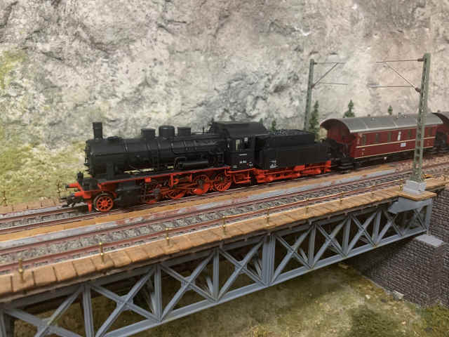 Historische Dampflokomotive der Baureihe 56 mit roten Personenwagen auf der Stahlfachwerkbrücke der Paradestrecke des MEC Gernsheim, vor einer felsigen Gebirgskulisse.