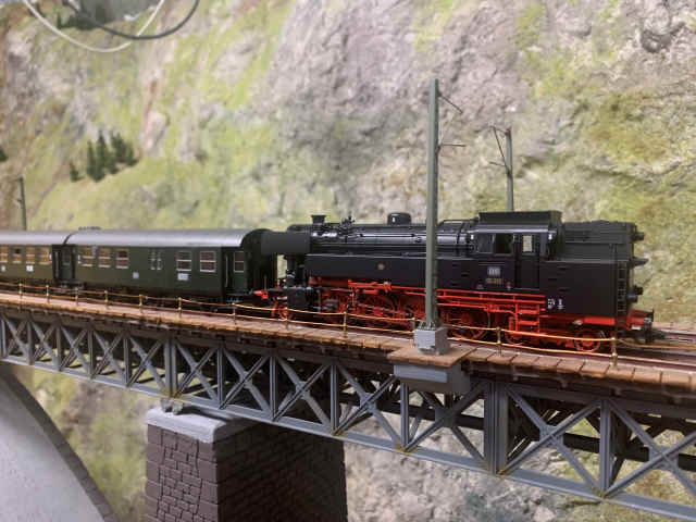 Historische Dampflokomotive der Baureihe 65 mit grünem Personenwagen auf der Stahlfachwerkbrücke der Paradestrecke des MEC Gernsheim, vor einer felsigen Gebirgskulisse.