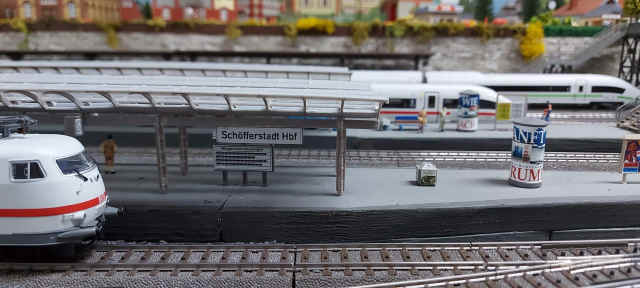 Weiße Elektrolokomotive der Baureihe 103 am Bahnsteig des Modellbahnhofs Schöfferstadt Hbf. Im Hintergrund steht ein ICE.