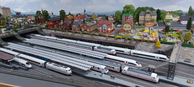Gesamtansicht des Modellbahnhofs Schöfferstadt Hbf mit mehreren weißen ICE- und Intercity-Zügen auf den Gleisen. Oberhalb des Bahnhofs liegt die detailreich gestaltete Modellstadt.