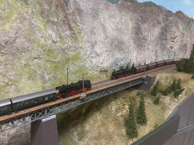 Zwei historische Dampflokomotiven der Baureihen 65 und 56 begegnen sich mit ihren Zügen auf der Brücke der Paradestrecke des MEC Gernsheim vor einer felsigen Gebirgskulisse.
