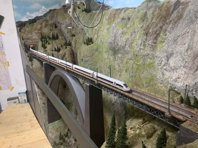ICE 4 auf einer hohen Eisenbahnbrücke vor einer felsigen Gebirgslandschaft auf der Modellbahnanlage des MEC Gernsheim.