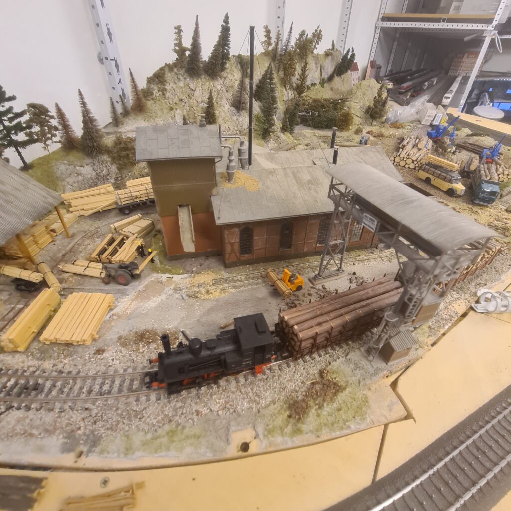Nachgebildeter Holzverladeplatz mit kleinen Betriebsgebäuden, Baumstämmen und einer Dampflokomotive.